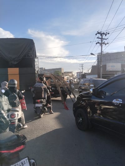 In Blangpeuria, Indonesia, on a sunny day with scattered clouds, traffic moves along a paved road. Several motorcycles are present, with one in the foreground carrying a person with a black backpack and wearing a helmet. Behind it, another motorcycle carries two individuals, one wearing a helmet and a backpack, and another passenger. A third motorcycle with two passengers is further back.
A dark-colored pickup truck is stopped on the right side of the road, loaded with metal rods. To the right of the truck, a black SUV is visible, and partially obscuring it is a black vehicle in the foreground, likely a car.
Buildings line the street, including one with a sign reading "Agen BSI". Electrical poles and wires are visible overhead, indicating urban infrastructure. The scene appears to capture a moment in the daily commute or transportation of goods in this Indonesian city.