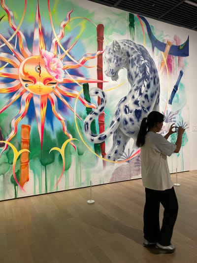 A person, viewed from the back, is standing in front of a large, colorful mural in what appears to be a museum or gallery. The mural depicts a stylized sun with a face, a snow leopard, bamboo stalks, and abstract elements. The person is holding up a mobile phone and taking a photograph of the artwork. The floor is made of polished wood, and stanchions are visible in the foreground, suggesting a public space. The overall impression is one of appreciation for art in a contemporary setting. The scene is located in Jinniu District, China.