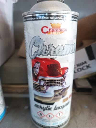 A close-up, slightly dusty shot focuses on a can of Champion color Chrome acrylic lacquer spray paint. The can features a graphic of a classic red car with chrome accents. The background is blurred, but appears to be a storage area, possibly a workshop or garage, with shelves and other objects visible. No people or animals are present. The specific location within Durlești, Moldova is not discernible from the image.