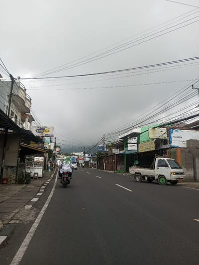The image presents an eye-level view of a bustling street in what appears to be a town or village under a heavily overcast, grey sky. The asphalt road, marked with a solid white line on the left shoulder and a dashed white line in the center, stretches into the distance.
On the left side of the street, a person riding a black motorcycle away from the camera is visible in the middle ground. The rider is wearing a white and black patterned jacket and a distinctive bright green head covering. Adjacent to the road is a paved sidewalk, along which a row of buildings stands. In the foreground, there's a multi-story building with balconies, and further down, various commercial establishments and stalls are visible. One prominent sign on the left reads "heny pod," and a larger sign above it clearly displays "SAMSUNG." Another sign below "heny pod" shows partial text like "Dapur..." and "Coffee." A street vendor stall with food items is also visible on the left.
On the right side of the street, a small white pickup truck (ute) is parked, facing away from the camera. Its front two wheels are fitted with striking bright green hubcaps or rims. Behind the truck, a series of single-story commercial buildings line the road, some featuring green facades or awnings. A pile of grey concrete blocks is stacked beside one of these buildings on the far right. Several signs are visible on this side, including "APOTEK" (Pharmacy) and "TB. SUJIYA."
A dense network of black power lines and communication cables crisscrosses high above the street, a prominent feature of the overhead space. The overall lighting is soft and diffused due to the overcast conditions. In the far distance, beyond the buildings, the faint silhouettes of hills or mountains are discernible through the atmospheric haze. The scene depicts an active, everyday environment with various vehicles and open businesses, characteristic of urban or semi-urban areas.