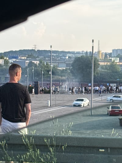 The image captures a scene in an outdoor urban environment, possibly a parking lot or an open area, with a crowd of people and several vehicles. In the foreground, a man with a shaved head, wearing a black t-shirt and white shorts, is seen from the back, looking towards the event. The background reveals a hazy atmosphere, suggesting either smoke or dust, and a gathering of individuals.

Further in the background, there are several cars, including a white sedan and a red sedan, parked or driving on a paved surface. The environment is characterized by a mix of industrial and green elements, with buildings, trees, and what appear to be utility poles visible in the distance. The sky is bright and appears to be daytime.

The setting is identified as the vicinity of the AmberSky shopping center in Gdańsk, Poland. This location is known for its urban landscape, featuring commercial buildings, infrastructure, and open spaces, consistent with the visual elements present in the photograph.