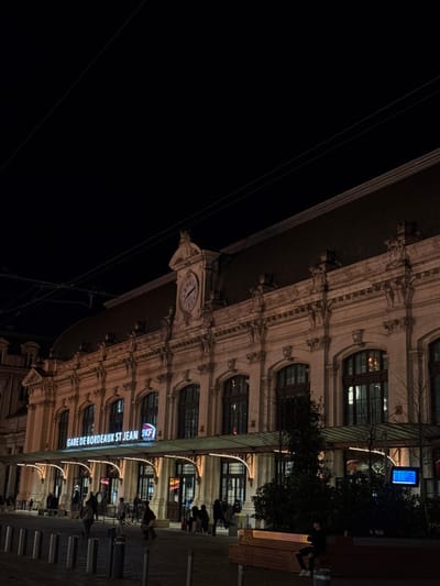 The image displays the exterior of the Gare de Bordeaux Saint-Jean railway station in Bordeaux, France, at night. The station's grand, light-colored stone facade is prominent, featuring classical architectural elements, numerous arched windows, and a central clock face on the upper level. Below the clock, an illuminated sign reads "GARE DE BORDEAUX ST JEAN" next to the SNCF logo. A wide, green-tinted canopy with integrated lighting extends over the main entrance area.

In the foreground, a paved pedestrian zone is present, lined with dark bollards. Numerous individuals are visible across this area, generally walking towards or away from the station entrances; several people near the entrance possess luggage. One person is seated on a long, light-brown wooden bench in the lower right quadrant. Dark shrubbery and bare tree branches are positioned in the midground. A blue-illuminated digital information screen is partially obscured by foliage on the right. The station facade and entrance area are brightly illuminated by warm artificial lighting, contrasting with the dark night sky, which contains visible overhead wires.