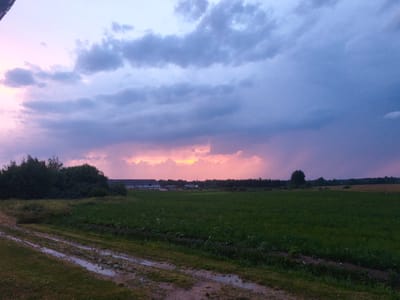 This is a landscape photograph taken at dusk in Kyviškės, Lithuania. The scene depicts a rural setting with a large, dark green field in the foreground, bordered by a muddy track with puddles. In the middle ground, a line of dark green trees separates the field from a cluster of buildings, possibly a farm or industrial complex, which are lighter in color. A dense forest borders the horizon.
The sky is dramatic, with heavy clouds tinged with shades of purple, blue, and pink, indicating the setting sun. There is a break in the clouds through which a bright, warm orange light is visible, suggesting a sunset or a distant storm illuminated by the sun. The overall mood is serene and somewhat melancholic, with the fading light and vastness of the landscape contributing to this feeling. The weather appears to be unsettled, with the dark clouds suggesting a possibility of rain. No people or animals are visible in the image, and there is no visible text.