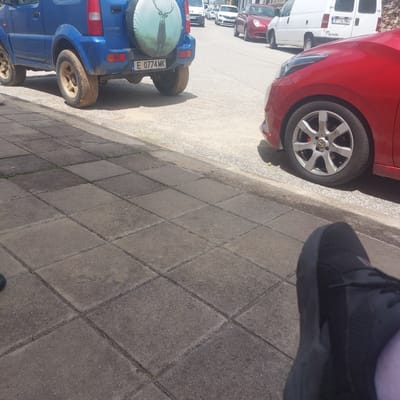 The image depicts a street scene in Ablanitsa, Bulgaria, focusing on the rear of a blue Suzuki Jimny and the front of a red car. The Suzuki Jimny, with license plate E 0774 MK, is parked on the right side of the frame. It has a spare tire mounted on the back with a cover featuring a deer graphic. The red car, a hatchback, is partially visible on the right, with its front wheel and fender in the foreground. The street is paved with asphalt and bordered by a sidewalk made of square concrete tiles. In the background, other vehicles, including a white van and a red sedan, are visible parked or driving on the street, suggesting a typical urban environment. The lighting suggests it is daytime, likely sunny, with shadows cast on the ground. The image appears to be taken from a low angle, possibly from a person sitting or crouching on the sidewalk.