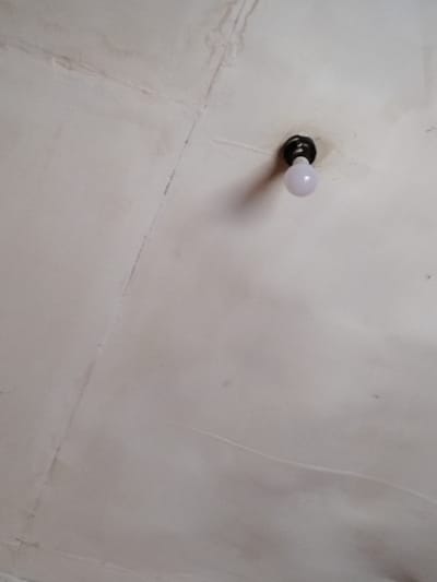 An upward-angle photograph depicts an interior ceiling surface in Muka, Indonesia. A single, spherical, opaque white light bulb is affixed to a dark, circular ceiling-mounted fixture, positioned in the upper-right quadrant of the frame. The ceiling is a light, possibly off-white, color and displays extensive discoloration, dark streaks, and irregular blotches consistent with moisture damage or mold. Visible seams and hairline cracks are present across the ceiling surface, particularly prominent in the upper-left and central portions. No individuals, actions, or interactions are visible. The light bulb appears unlit.