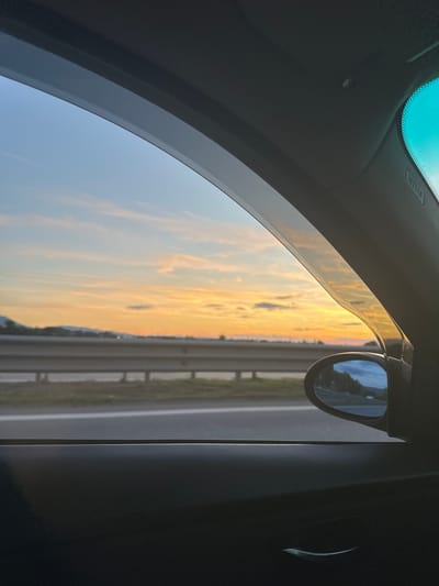 The image, taken from inside a car, shows a sunset view through the passenger window. The sky is a gradient of blue, orange, and yellow, with scattered clouds. A highway barrier and trees are visible in the distance. A side mirror reflects a portion of the scenery. The interior of the car, including the door panel and window trim, is dark and out of focus. The location is identified as Krivina, Bulgaria.