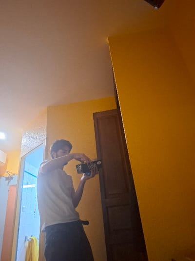 A person holds up a phone while standing in a brightly lit bathroom. The person is wearing a white t-shirt and black pants. The bathroom features a large window, blue and white tiles, and yellow towels. Above the person is a wooden cabinet against a yellow wall. The rest of the room consists of white and yellow walls and ceilings. The image is rotated.