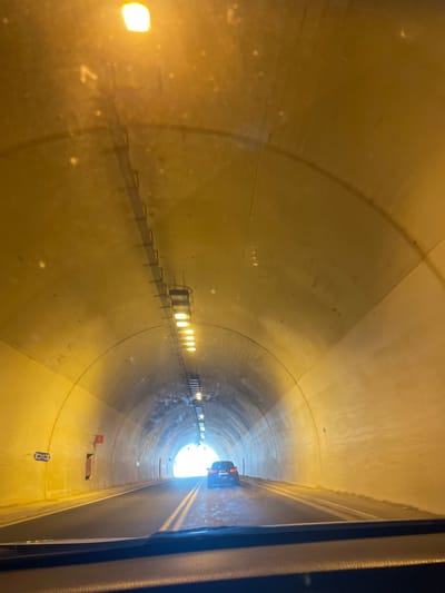The image shows the interior of a vehicle looking out towards the end of a tunnel. The tunnel walls are illuminated by overhead lights, creating a yellow glow. A single car is visible ahead, driving towards the bright exit of the tunnel. There are lane markings on the road and some signage on the left wall of the tunnel. The overall scene depicts travel through an underground passage.