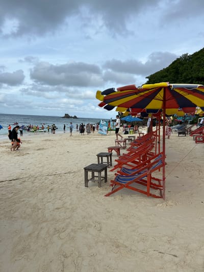 The image shows a crowded beach in Phru Nai, Thailand. The sky is overcast with dramatic clouds. In the foreground, several colorful beach chairs and umbrellas are set up on the sand. A row of red beach chairs with blue and yellow striped cushions is lined up under a large, multi-colored umbrella. Small wooden tables are placed between some of the chairs. Further back, numerous people are visible on the beach and in the shallow water. Some are standing, while others are interacting with jet skis, suggesting recreational activities. The water is calm, and a rocky island formation is visible in the distance. The sand appears light-colored and textured with footprints.