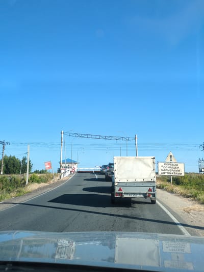 The image shows a view from inside a vehicle looking forward on a two-lane asphalt road. The sky is clear and blue. On the right side of the road, there is a white sign with black text that reads "ПЕРЕЕЗД ОСНАЩЕН БАРЬЕРАМИ - АВТОМАТАМИ" (Crossing equipped with barriers - automatons), and above it, a triangular warning sign with an exclamation mark. Further ahead, a railway crossing is visible with barriers that are partially lowered. There is a small building with a blue roof next to the crossing. Several vehicles are on the road, including a white trailer in the foreground and other cars behind it, all appearing to be stopped or moving slowly. Power lines run overhead across the road. The surrounding environment is rural with dry grass and some green trees on the sides of the road. The scene is likely a railway crossing in the city of Dzerzhinsk, Russia, as indicated by the text on the sign.