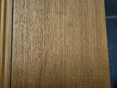 The image displays a close-up view of a wooden surface with a prominent grain pattern. The wood is a warm, medium brown color with darker vertical lines representing the grain. On the left side of the image, there is a distinct vertical edge, suggesting a corner or the side of a panel. This edge has a slightly lighter tone and appears to have some subtle vertical ridges. The right side of the image shows a transition to a dark, out-of-focus background, which is likely a solid color such as black or dark blue. The lighting appears to be coming from the left, casting subtle shadows that highlight the texture of the wood grain. There are no people, animals, or any other discernible objects or actions in the scene. The image is a still shot and provides no information about the location in Bugulma, Russia, or any forensic details beyond the visual characteristics of the wood.