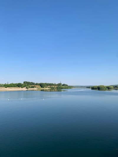 The image depicts a wide, calm body of water, likely a river or lake, with a clear blue sky. On the left side of the frame, the sky dominates, a serene expanse of blue. The right side of the frame is occupied by the water, which reflects the sky and the green, tree-lined shore.
The shore is densely covered with lush green trees, creating a natural border between the land and the water. The trees appear healthy and vibrant, suggesting a warm season, likely summer. The water is relatively still, with gentle ripples visible, and it reflects the colors of the sky and the vegetation on the shore. Several white buoys or markers are scattered across the water, indicating a navigable channel or designated area. There are also a few small, dark specks in the water, which could be birds or other aquatic life.
The overall scene is peaceful and tranquil, conveying a sense of natural beauty and serenity. The time of day appears to be daytime, given the bright blue sky and the clear visibility. The weather seems pleasant, with no clouds visible in the sky. While no specific text is visible in the image, the natural landscape suggests an outdoor recreational area or a scenic route. The location context of Ustia, Moldova, places this scene in a specific geographical region.