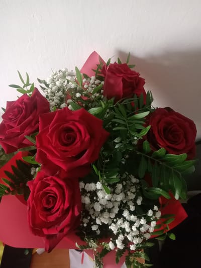 A bouquet of red roses and white baby's breath is presented against a white textured wall. The bouquet is wrapped in bright red paper. The lighting suggests an indoor setting, likely a home or commercial space. There is a subtle shadow cast on the wall behind the bouquet, indicating a light source from the right. The image does not contain any identifiable landmarks or clues to determine its location in Kielce, Poland. The scene is static and appears to be a still life photograph of the floral arrangement.