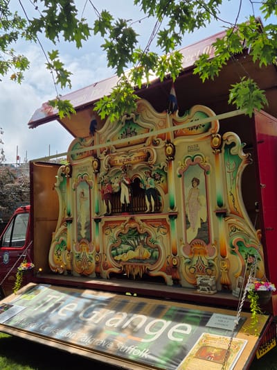 The image depicts a street organ, known as a dance organ, displayed in the back of a red truck. The organ is elaborately decorated with colorful paintings of figures and landscapes. On the opened tailgate of the truck, a large banner with "The Grange Musical Collection Palgrave Suffolk" is displayed, indicating the location of the organ. The scene appears to be outdoors, with trees and a cloudy sky visible in the background. There are no people visible in the image. The organ has various decorative panels and some small figures appear to be part of its mechanism. A small basket with flowers hangs from a chain to the left of the organ. Additional signage, including information about CDs for sale and an "Open Days 2026" flyer, is also visible on the tailgate. The overall impression is of a mobile exhibition of a musical instrument.