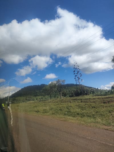 The image captures a scene in Kibirichia ward, Kenya. A dirt road stretches into the distance, bordered by grassy terrain that slopes gently upwards. A hill covered in trees and vegetation occupies the center background. Scattered trees and taller plants are visible along the hillside. Overhead, a sky with both clear blue patches and numerous puffy white clouds is crisscrossed by power lines. At the left edge of the road, two people are standing. One is wearing a yellow safety vest. The scene appears to be shot from inside a vehicle, with a slight reflection visible on the lower left of the image.