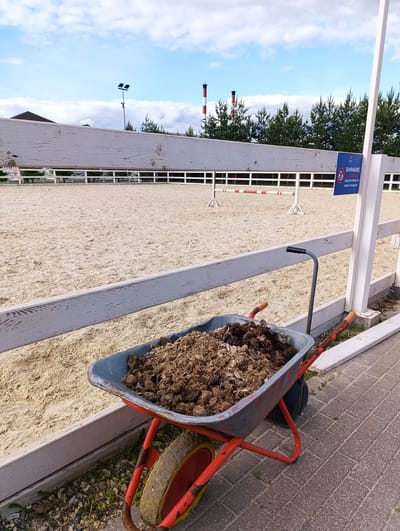 The image depicts a wheelbarrow filled with manure. The wheelbarrow is situated next to a white fence enclosing a sand arena. In the background, there are trees and industrial chimneys. A blue sign with Cyrillic writing is attached to the fence. The wheelbarrow is worn with red frame. The overall scene appears to be an outdoor equestrian facility. The location is the city of Chernogolovka, Russia.