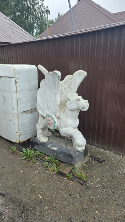 The image shows a white concrete sculpture of a winged creature, possibly a griffin or a dragon, situated outdoors. The sculpture is detailed with visible wings, a head with a snout, and clawed feet. A circular emblem with a floral design, featuring red petals and a green border, is attached to the creature's body. The sculpture rests on a dark, irregular-shaped base.

The setting appears to be an outdoor area with a brown corrugated metal wall in the background, suggesting a building or structure. The ground is a mix of gravel, dirt, and scattered debris, with some green weeds growing around the base of the sculpture and near some dark metal elements protruding from the ground. To the left of the sculpture, a portion of a silver-colored rectangular object, possibly a container or a piece of equipment, is visible. Further to the left, a sloping brown roof and a light fixture mounted on a pole can be seen, with trees and an overcast sky in the distance.

The lighting suggests it is daytime, and the wet appearance of the ground and the overcast sky might indicate recent rain or damp weather. There is no visible text in the image. The overall impression is one of a slightly weathered, possibly rustic, outdoor space.