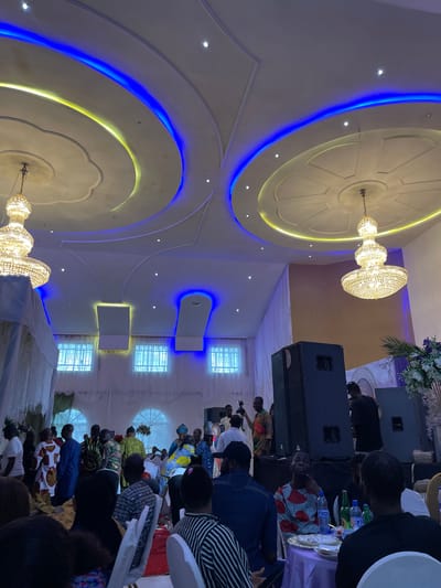 The image depicts an indoor event, possibly a reception or celebration, at a venue in Edopa, Nigeria. The room features a decorative ceiling with recessed lighting in blue and yellow, and two ornate chandeliers hang from the ceiling. A group of people are gathered, some standing and some seated at tables draped with purple tablecloths. Several individuals are dressed in traditional Nigerian attire. In the background, there are large speakers, suggesting a performance or DJ setup, and floral decorations. The windows in the background are covered by white drapery. The overall atmosphere appears festive.