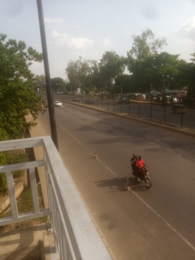 The image shows a street scene in Unguwan Dosa, Nigeria. The foreground features a white metal railing, suggesting a raised viewpoint, possibly a bridge or an elevated walkway. Beyond the railing, a paved road stretches into the distance, lined with trees and a dark fence. The sky is overcast, indicating it could be morning or late afternoon, with soft, diffused light.

The main activity visible is on the road where two people are riding a motorcycle. The rider, wearing a red shirt, and a passenger are moving away from the viewer. Their presence suggests everyday transportation. In the background, a white car is driving on the road, and further down, a yellow three-wheeled vehicle, possibly a tuk-tuk, is visible. There are also buildings and trees on the other side of the road, hinting at an urban or semi-urban environment. A small potted plant sits on the side of the road near the motorcycle, a small detail adding to the scene's ordinariness. No visible text is present in the image. The overall impression is a calm, everyday street scene in a populated area.