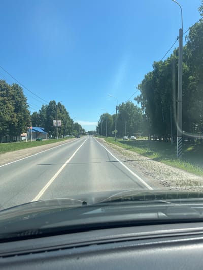 A first-person perspective shot from a car shows a straight, paved two-lane road under a clear blue sky. Trees line both sides of the road, with some buildings visible on the left. Streetlights are present along the road. The car's dashboard is visible in the foreground. The location is identified as Yadrin, Russia.
