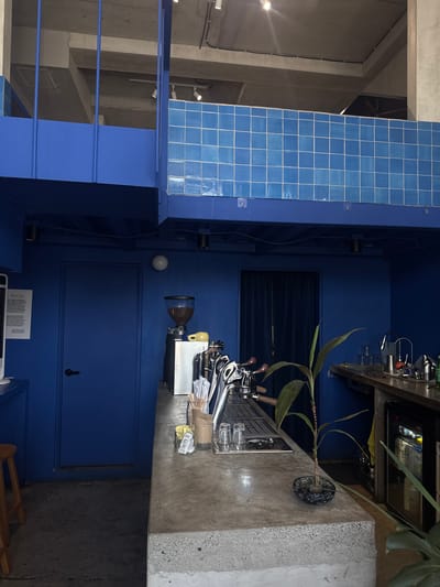 The image depicts the interior of a coffee shop with a prominent blue color scheme, located in San Pedro Garza García, Mexico. The scene is viewed from a slightly elevated angle, looking across a concrete counter. On the left, a blue door is visible, with a sign on the wall next to it. A coffee grinder sits on the counter to the right of the door. The counter itself is a textured concrete surface, and on it are various coffee-making implements, cups, and a small potted plant. In the background, a dark blue curtain hangs, obscuring what is behind it. To the right of the counter, a stainless steel espresso machine is partially visible, and further to the right, a commercial refrigerator with glass doors is present. The upper portion of the image shows exposed concrete ceilings and blue tiled walls, suggesting an industrial or modern architectural style. There are no people visible in the frame.