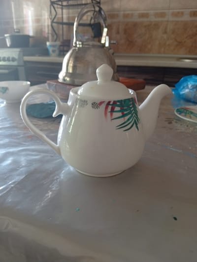 In a brightly lit kitchen in Chigatay, Uzbekistan, a white ceramic teapot with a decorative design of green leaves and red accents sits on a transparent plastic tablecloth. The teapot has a curved handle and a spout. In the background, a silver kettle is visible on a shelf, along with other kitchen appliances and tiled walls. The scene suggests a domestic setting, possibly a mealtime or a gathering.