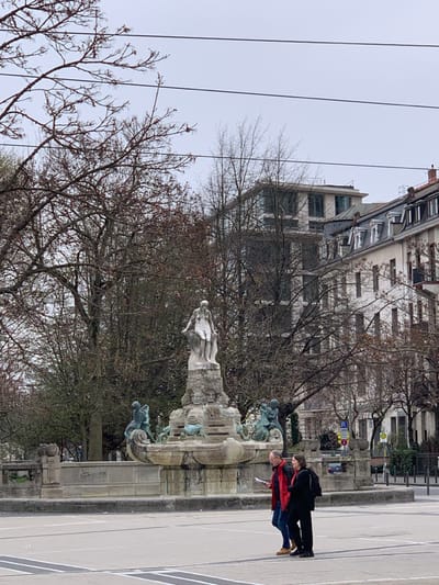 A stone fountain is centered in a public square in Frankfurt am Main, Germany, located at latitude 50.109 and longitude 8.673. The fountain features a central statue of a woman in a flowing robe, standing on a pedestal, with water cascading down the sides. To the left and right of the central statue are two statues of smaller figures. The fountain is surrounded by a low stone wall. In the background, there are trees, a building, and street signs. A couple is standing in front of the fountain. The man is looking at a map, and the woman is holding a backpack. The ground is a gray concrete. The sky is overcast. The trees are mostly bare, with some green leaves.