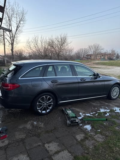 A dark grey Mercedes-Benz C-Class (W205) station wagon is parked outdoors on a muddy, partially paved surface with patches of melting snow. The vehicle's exterior appears recently cleaned. A green and black hydraulic floor jack lies on the ground near the passenger side, with a green metal handle/bar extending from it. A red and black cordless impact wrench or drill is positioned on the ground to the rear left of the vehicle. The immediate foreground consists of damp soil and grey, textured paving stones. In the background, numerous deciduous trees with bare branches are visible against a light blue sky with faint orange hues near the horizon, indicative of either sunrise or sunset. Overhead, multiple power lines traverse the frame. Further in the distance, a field of dry grass and several weathered, low-rise residential structures with tiled roofs are discernible, characteristic of a rural or semi-urban setting in Bohot, Bulgaria. No individuals are present in the scene.