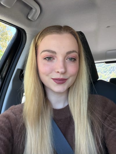 The image features a young woman inside a car, likely in Sant'Alessio Siculo, Italy, captured during the daytime. She has long, straight, blonde hair that falls over her shoulders and part of the car seat. Her complexion is fair, and she has green or hazel eyes enhanced with subtle makeup, including eyeshadow, mascara, and a muted reddish-pink lipstick. She wears a brown, soft-textured sweater or top, and a blue-grey seatbelt is visible across her chest.

The woman is looking directly at the camera with a gentle, relaxed smile, suggesting a positive and calm mood. The setting is the interior of a vehicle, with a grey headliner and a matching overhead grab handle visible in the background. Through the car window, vibrant green foliage and natural light indicate a bright, clear day outdoors. No visible text is present in the image.