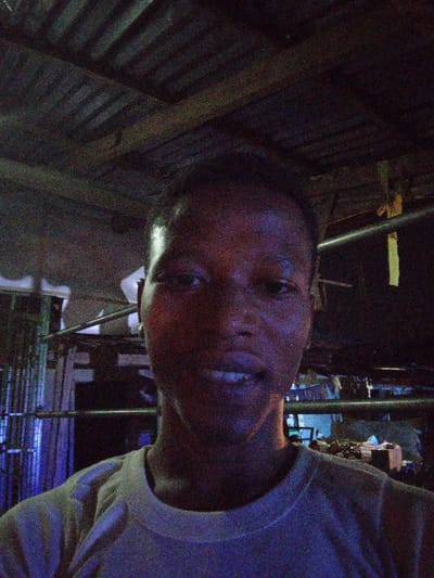 A person with short hair wears a light-colored t-shirt. The background is dimly lit and appears cluttered. A partially visible metal structure stands behind the person. The ceiling is made of planks or boards. The image has significant noise and low contrast.