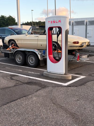 A cream-colored convertible Alfa Romeo Spider is parked on a trailer at a Tesla charging station in Hogstorp, Sweden. The car is facing right, with its front on the left side of the frame. The Tesla charging station is white with red accents and the Tesla logo is visible. The car is being towed by a trailer with two wheels on each side. The trailer is parked in a marked parking spot. The background shows some trees and a building. The sky is partly cloudy. The license plate of the car is V A 1751. The license plate on the trailer is VA 1456.