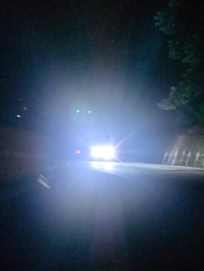 The image depicts a nighttime scene on a road, likely in or around Kulang, India, due to its mountainous terrain suggested by the presence of a retaining wall on the right side of the road and foliage. The primary focus is a vehicle approaching, its headlights illuminating the scene with intense brightness, causing significant glare and obscuring details. The road appears to be paved and curves to the left. The surroundings are dark, with trees visible on the right and hints of foliage on the left. The overall impression is one of driving at night in a rural or semi-rural mountainous area. No people are clearly visible due to the lighting conditions and the focus on the approaching vehicle.