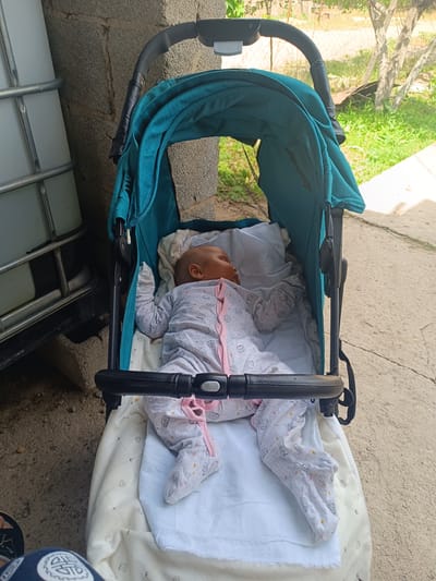 The image shows a newborn baby sleeping in a teal-colored stroller. The baby is wearing a light gray onesie with a pattern of small animals and pink trim. The stroller is on a concrete surface, with a building and some greenery in the background. The location is identified as Shamalgan, Kazakhstan.
