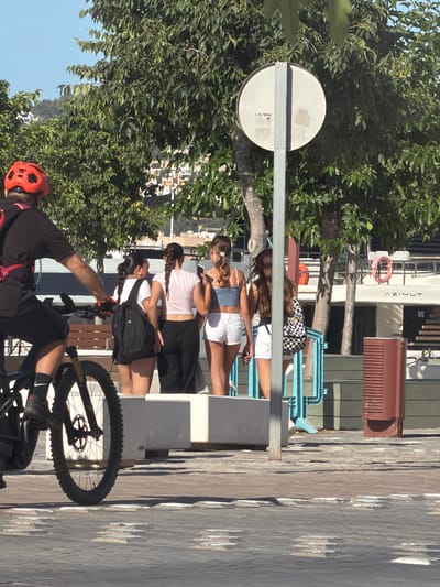 The image depicts a street scene in Eivissa, Spain, likely near the harbor, characterized by a clear blue sky and lush green trees. In the foreground, a person wearing a red helmet and black athletic clothing is riding a bicycle. Their right leg is on the pedal, and their left leg is extended downwards. The bicycle has a prominent front wheel and part of the frame visible.
Behind the cyclist, four young women are standing. The woman on the far left wears a black backpack and a white t-shirt. Next to her, a woman with dark braided hair wears a light pink crop top and black pants. To her right, another woman with light brown hair also braided, is wearing a light blue crop top and white shorts. The woman on the far right, with blonde hair, wears a light blue top and white shorts. One of the women is holding a smartphone.
In the background, a white boat with the name "AZIMUT" is docked. Several large green trees create a natural backdrop, and parts of buildings are visible in the distance. A grey signpost with a circular white sign stands prominently in the middle ground. The ground is paved with textured stones. A dark brown rectangular structure with ventilation slits is positioned to the right of the women.