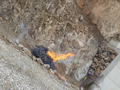 The image depicts a natural gas fire burning at the entrance of an old mine shaft in the city of Madan, Bulgaria. The fire is characterized by bright orange and yellow flames licking upwards from a pile of dark, twisted metal debris. Wisps of white smoke emanate from the fire and blend into the rocky background. The surrounding environment is rugged and appears to be a mountainside or excavation site. The terrain is composed of loose gravel, rocks of various sizes, and exposed rock faces with distinct geological strata. To the right of the fire, a concrete structure with a collection of stones is visible, suggesting ongoing construction or a boundary marker. The scene is captured from an elevated perspective, looking down into the mine entrance.