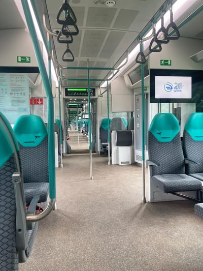 The image captures the interior of a modern train carriage, likely a commuter train, with a focus on the seating and aisle. The seats are upholstered in a patterned grey fabric with teal accents and feature the brand "ИВОЛГА" (IVOLGA) in white lettering. Overhead, triangular black handgrips are suspended from silver rails, which are also part of a teal-colored support structure running along the length of the carriage. A large screen on the right displays a logo with "20 лет ЦППК" (20 years TsPPK). Emergency exit signs with green pictograms are visible on the walls. The floor is carpeted with a speckled grey material. In the distance, the aisle extends, revealing more rows of seats and a few passengers, though they are not clearly discernible. The overall environment is clean, well-lit, and appears to be in operational condition. The reference to Sokolovo, Russia, is not visually derivable from the image itself.