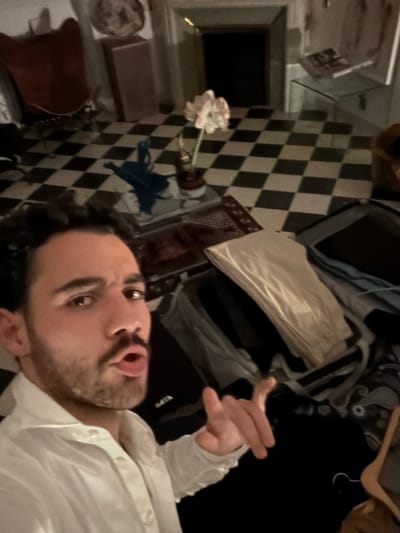 An indoor scene in Mauléon-d'Armagnac, France, depicts a dimly lit room with a black and white checkered floor. In the immediate foreground, a male individual, appearing to be in his twenties or thirties with dark hair and a beard, is visible from the chest up, wearing a white collared shirt. He is looking directly at the camera with his mouth slightly open in an expressive manner, and his right hand is raised and slightly curled in a gesture.
To the right of the subject, an open black suitcase containing various folded clothes, including a light-colored item on top, is present. Another smaller, dark bag is partially visible beside it. Behind the subject and suitcases, a square glass coffee table holds a brown pot with a single plant bearing white flowers and a small blue sculptural object. A patterned rug is visible underneath the table.
Further back in the room, a large fireplace with a dark, possibly empty, firebox is centered against a wall. To the left of the fireplace, a brown lounge chair with a metal frame is visible. To the right of the fireplace, a small glass side table holds an unidentified item. The lighting in the room is ambient and appears to be artificial, casting some shadows.