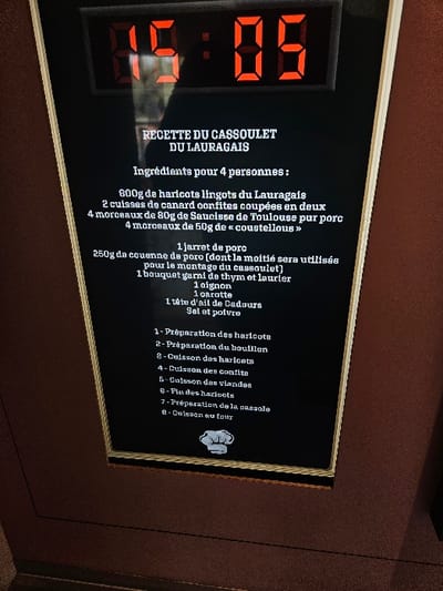 A vertical digital display, framed in dark brown with a golden inner border, is mounted on a dark reddish-brown wall in Avignonet-Lauragais, France. The screen's top section features a red digital clock illuminated to show "19:05". Below the clock, a white-on-black text field presents the title "RECETTE DU CASSOULET DU LAURAGAIS" (Recipe of Cassoulet from Lauragais). This is followed by a list of ingredients for four people, detailed in French, including Lauragais cannellini beans, duck confit legs, Toulouse sausage, various pork cuts (jarret, coustellous, couenne), a bouquet garni, onion, carrot, Cadours garlic, salt, and pepper. Beneath the ingredient list, eight numbered steps outline the preparation process in French, ranging from bean preparation to final oven baking. A white stylized chef's hat icon is positioned at the bottom of the recipe steps. A faint reflection is present on the display's surface.
