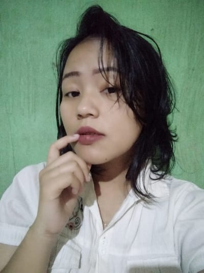 A young Asian woman poses indoors against a muted green background. She has dark hair and fair skin and wears a white button-down shirt. Her hair is styled with bangs and some layers. Her lips are colored with a matte berry shade. She holds a finger lightly to her lips in a thoughtful or pensive pose, looking slightly off-camera.