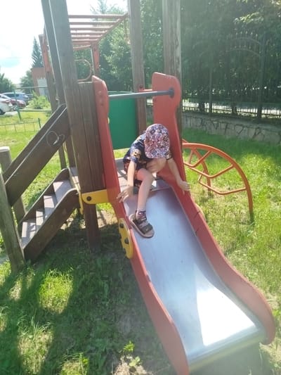 In Yalutorovsk, Russia, on a sunny day, a young child wearing a patterned cap and dark clothing is at the top of a metal slide on a wooden playground. The child is positioned to slide down, with their hands on the edge of the slide. The playground equipment is made of wood and red metal, with a set of wooden stairs leading up to a platform. In the background, trees and a black metal fence are visible. The ground is covered with green grass and some patches of dirt.