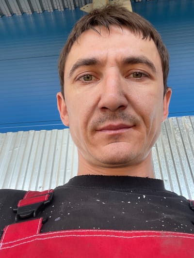 The image is a close-up, selfie-style, low-angle shot of a man wearing red and black overalls over a black shirt. He has short brown hair and brown eyes, and a light stubble on his face. The background consists of a corrugated metal wall and a blue ceiling or overhang. The overall setting appears to be outdoors, possibly in a work area or under a covered structure, with elements suggesting a construction or industrial environment. There are no other individuals or significant objects visible in the frame besides the man and his immediate surroundings. The lighting is natural and somewhat harsh, casting slight shadows. The image quality is clear, and details of the man's skin texture and the fabric of his clothing are discernible. The scene is devoid of any contextual clues that would place it specifically within the city of Сокуры, Russia, or any other urban location.