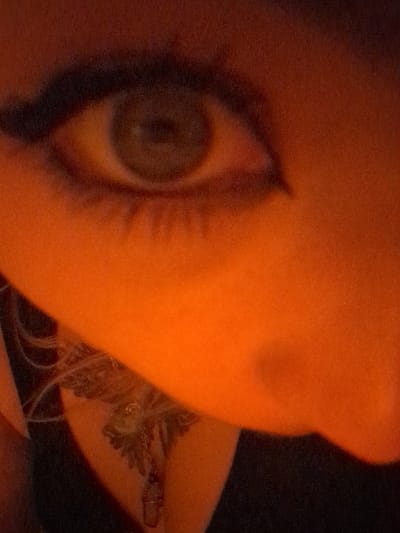 The image presents a close-up, high-angle view of a person's right eye, upper cheek, and a portion of the nose, bathed in intense orange-red ambient lighting. The open eye features a green/hazel iris, a dark pupil, and visible upper and lower eyelashes. Dark eyeliner defines the upper lid, extending into a winged form. The bridge and tip of the nose are partially visible on the right side of the frame. Below the eye, on the person's neck or upper chest, a dark-inked tattoo depicts a detailed winged creature, possibly a bird, with a chain featuring a small, translucent charm and a dark, cylindrical pendant visible draped over it. The background is dark and out of focus. The scene is situated in Area D (Elaho/Garibaldi), Canada.