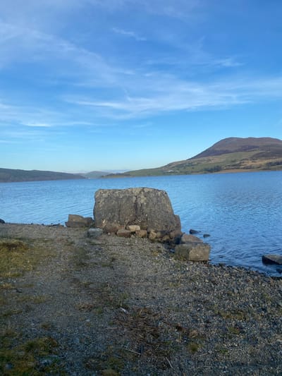 The image depicts a landscape scene in Bala, United Kingdom, showing a body of water, likely a lake, adjacent to a shoreline composed of small gravel and rocks. A large, grey rock sits at the water's edge, with smaller stones nearby. The water surface has a slight ripple effect. In the background, a mountain range is visible on the opposite side of the lake, with varying shades of brown and green indicating vegetation. The sky above is blue with wispy, scattered clouds.