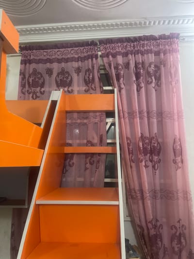 The image depicts a close-up view of the interior of a bedroom in Kporva, Nigeria. The primary subjects are an orange bunk bed ladder and a set of sheer pink curtains with a dark purple damask pattern. The bunk bed ladder is constructed from wood and painted a bright orange, with white trim. It features two visible steps, suggesting it leads to an upper sleeping area. The curtains are draped across a window, partially obscuring the view outside. The ceiling above is white with textured circular patterns. The overall impression is of a child's bedroom, possibly with a focus on functionality and vibrant color.