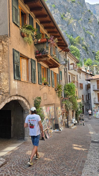 This media file depicts a person standing on a cobblestone street in what appears to be a European village. The scene is outdoors, with buildings lining the street and a steep, rocky mountainside visible in the background. The time of day seems to be daytime, with sunlight casting shadows on the cobblestones. The weather appears to be fair.

The main subject is a person, viewed from the back, wearing a white t-shirt with text and graphics, blue shorts, and sneakers. The person is standing near an arched entryway. In the foreground, a collection of paintings or artwork is laid out on the ground, suggesting a street market or an artist's display. The buildings have balconies with plants, and some windows have open shutters. The architecture suggests an older, European style.

The text on the t-shirt is partially visible. It appears to say "LAINÉSİM" and has colorful graphics.

The location context provided is Limone sul Garda, Italy, which aligns with the visual cues of the narrow street, the lakeside town architecture, and the proximity to mountains. The overall impression is one of a charming, scenic village setting, possibly during a tourist season.