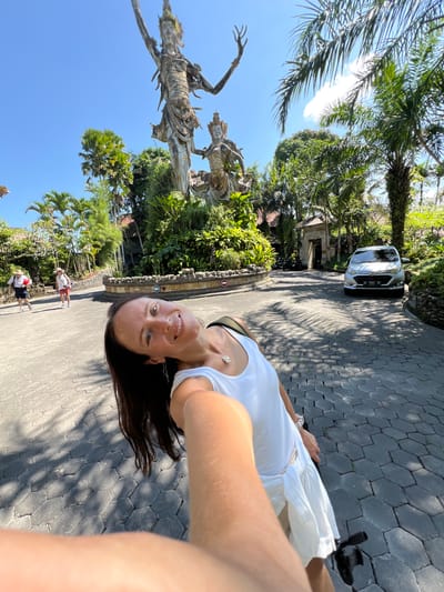 A selfie taken at the entrance to the Royal Pita Maha Resort in Kedewatan, Indonesia. A woman in a white top stands on a gray stone path, smiling at the camera. Behind her, a white car is parked in the middle of the path, flanked by greenery and trees. To the left stands a large sculpture. Further behind the woman, two people walk on the path. The sky is blue.