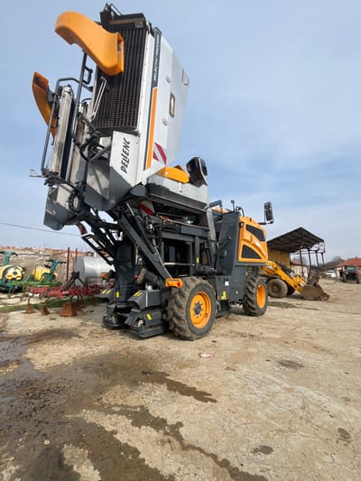 The scene depicts an outdoor agricultural machinery storage or maintenance yard in the city of Terziysko, Bulgaria, under a partially cloudy blue sky.

The primary subject is a large, self-propelled Pellenc grape harvester, predominantly grey, orange, and black, positioned centrally. The machine's harvesting head is elevated, exposing vertical internal rollers, and displays "PELEENC" and "SELECTIV PROCESS" branding. It features large, multi-lugged black tires with orange hubs.

To the right, a yellow JCB backhoe loader or wheel loader, equipped with a front bucket, is parked beneath an open-sided shed with a corrugated metal roof. A red tractor is partially visible further to the right.

To the left of the grape harvester, various agricultural implements are visible, including green and yellow spraying equipment and a red plow attachment. Behind these, a large, horizontal, silver cylindrical tank is present.

The ground is an unpaved surface of dirt and gravel, marked with several dark, irregular stains consistent with pooled water or oil. No individuals are visible within the frame; all machinery appears stationary.