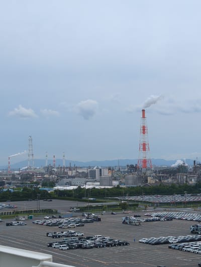 This is an aerial, eye-level shot of a large industrial area in Yokkaichi, Japan, on an overcast day. In the background, a sprawling complex of factories, pipes, and towers are visible. Smoke billows from several smokestacks, including a prominent red and white striped one. In the mid-ground, a vast parking lot is filled with thousands of cars, likely awaiting shipment. A few vehicles, including a car carrier truck and a forklift with a person on it, are in motion. The foreground shows more of the expansive parking lot. The overall scene conveys a sense of large-scale industry and commerce.