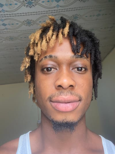 The media file is a close-up, head-and-shoulders selfie of a young Black man with dreadlocks. His hair is dark at the top, transitioning to a blonde hue at the ends. He has a light beard and mustache. His eyes are open, and his lips are slightly parted. He is wearing a white tank top. The background appears to be a wall with patterned wallpaper. The lighting suggests it is daytime, possibly indoors. The image is oriented sideways. There is no visible text. The context provided indicates the location is Port-Harcourt, Nigeria.