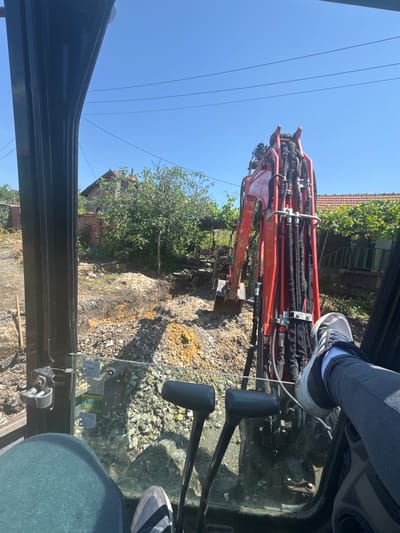 The image captures a view from inside the cabin of an excavator or similar heavy machinery, looking out over a construction or excavation site in a rural or semi-rural setting.
In the foreground, the lower body of the machine operator is visible, with legs clad in dark gray pants and athletic shoes (one grey Adidas shoe with white sole is prominent), positioned near the machine's control levers or pedals. Through the cabin window, the ground below appears disturbed, consisting of gravel, earth, and piles of dug-up soil, indicating ongoing work.
The midground is dominated by the robust, red arm of the excavator, equipped with numerous black hydraulic hoses secured by clamps. This arm extends into the work area, which features more disturbed ground, some green trees, and bushes. In the background, beyond the immediate work zone, traditional-style houses with reddish-brown tiled roofs are visible, nestled among greenery.
The upper portion of the image reveals a clear, bright blue sky traversed by several parallel power lines. The strong sunlight and lack of clouds indicate a sunny day, likely mid-morning or afternoon. Some small, unreadable text or logos are present on stickers affixed to the cabin window, one possibly containing "3X" or "13X".
The scene depicts an active earthmoving or construction operation, consistent with a worksite in Konstantinovo, Bulgaria, under clear weather conditions.